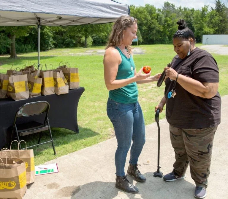 two women stand outdoors at a community food distribution table, looking at fresh vegetables with bags of food nearby