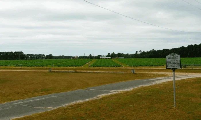 rural landscape with green crop fields on both sides of a dirt road leading to farm buildings in the distance. A historical marker for East Carolina Indian School stands on the right side under an overcast sky