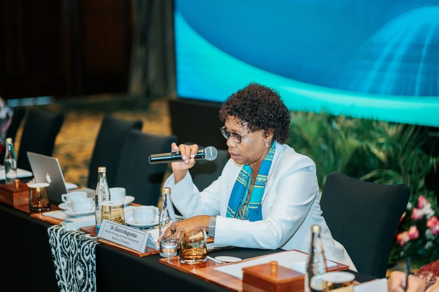 woman in a white blazer and blue patterned scarf speaking into a microphone at a conference panel table