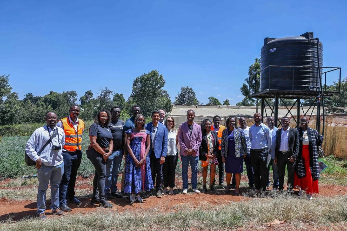 a group of visitors and staff stand together for a photo at an agricultural site, with fields and a water tank in the background